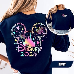 Puede incluir: Sudadera azul marino con un dise&ntilde;o colorido de Disney que presenta un castillo, fuegos artificiales y orejas de Mickey Mouse. El texto "Disney 2026" est&aacute; en degradado arco&iacute;ris. Una versi&oacute;n m&aacute;s peque&ntilde;a del dise&ntilde;o est&aacute; en el pecho izquierdo.