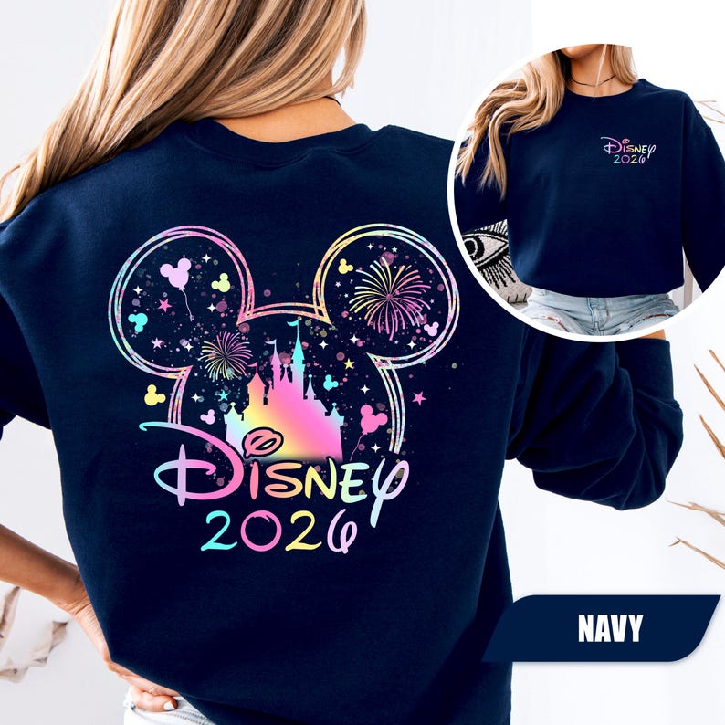 Puede incluir: Sudadera azul marino con un dise&ntilde;o colorido de Disney que presenta un castillo, fuegos artificiales y orejas de Mickey Mouse. El texto "Disney 2026" est&aacute; en degradado arco&iacute;ris. Una versi&oacute;n m&aacute;s peque&ntilde;a del dise&ntilde;o est&aacute; en el pecho izquierdo.