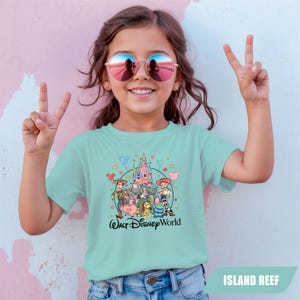 Puede incluir: Una camiseta azul claro con un gr&aacute;fico de los personajes de Toy Story de Disney y un castillo, con el texto "Walt Disney World". La camiseta la lleva una ni&ntilde;a peque&ntilde;a que lleva gafas de sol y hace el signo de la paz.