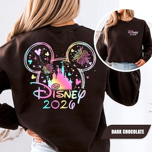 Puede incluir: Sudadera de color chocolate oscuro con un dise&ntilde;o colorido de Disney. El dise&ntilde;o incluye una silueta de Mickey Mouse con fuegos artificiales, un castillo y el texto "Disney 2026". Una versi&oacute;n m&aacute;s peque&ntilde;a del dise&ntilde;o est&aacute; en el frente.