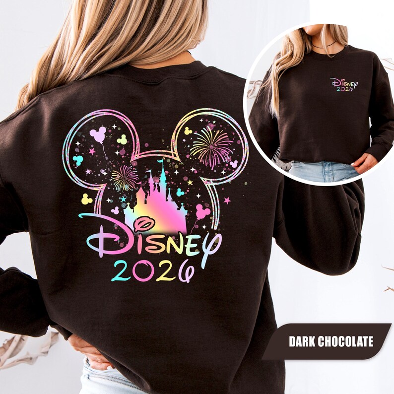 Puede incluir: Sudadera de color chocolate oscuro con un dise&ntilde;o colorido de Disney. El dise&ntilde;o incluye una silueta de Mickey Mouse con fuegos artificiales, un castillo y el texto "Disney 2026". Una versi&oacute;n m&aacute;s peque&ntilde;a del dise&ntilde;o est&aacute; en el frente.