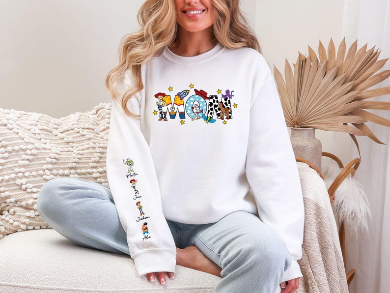 Op de afbeelding: Witte sweatshirt met het woord "Mom" in kleurrijke, cartoonachtige letters. De letters bevatten personages en afbeeldingen uit een populaire animatiefilm. De linkermouw heeft karakterillustraties en namen.