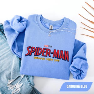 Puede incluir: Sudadera azul Carolina de Spider-Man con el logotipo de Marvel Studios. El gr&aacute;fico presenta las palabras "SPIDER-MAN" en rojo y "BRAND NEW DAY" en amarillo. Un collar plateado est&aacute; drapeado en el escote. La sudadera se combina con vaqueros azules.