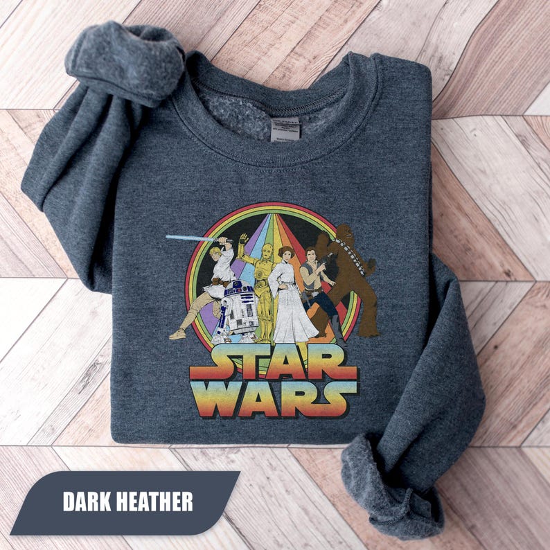 May include: Dark heather gray sweatshirt featuring a colorful Star Wars graphic. The graphic includes characters like Luke Skywalker, Princess Leia, and Chewbacca, with the words "STAR WARS" in a rainbow font. The text "DARK HEATHER" is at the bottom.