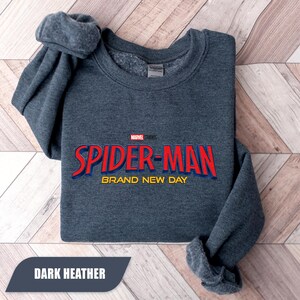 Puede incluir: Sudadera gris jaspeado oscuro con el logotipo de Spider-Man en rojo y azul y el texto "BRAND NEW DAY". El logotipo de Marvel Studios est&aacute; encima del texto de Spider-Man. La sudadera est&aacute; doblada sobre una superficie de madera clara.