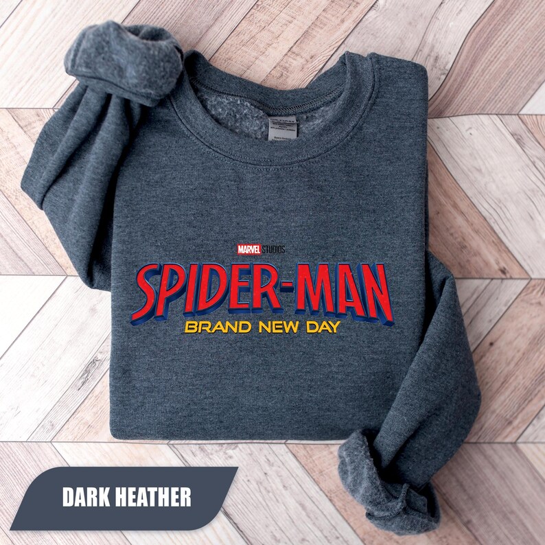 Puede incluir: Sudadera gris jaspeado oscuro con el logotipo de Spider-Man en rojo y azul y el texto "BRAND NEW DAY". El logotipo de Marvel Studios est&aacute; encima del texto de Spider-Man. La sudadera est&aacute; doblada sobre una superficie de madera clara.