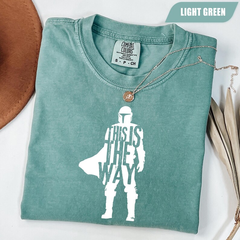 May include: A light green short-sleeve t-shirt featuring a white graphic of a helmeted figure in armor with the text "THIS IS THE WAY" integrated into the silhouette. The shirt has a crew neckline and a Comfort Colors brand label at the collar.