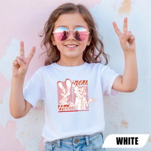 Puede incluir: Camiseta blanca con un gr&aacute;fico rojo de dos personajes de dibujos animados, un conejo y un zorro, con el texto "Wilds" y "Hopps Undercover". La camiseta la lleva una persona joven que lleva gafas de sol y hace el signo de la paz.