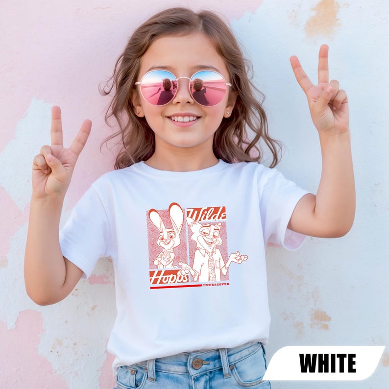 Puede incluir: Camiseta blanca con un gr&aacute;fico rojo de dos personajes de dibujos animados, un conejo y un zorro, con el texto "Wilds" y "Hopps Undercover". La camiseta la lleva una persona joven que lleva gafas de sol y hace el signo de la paz.