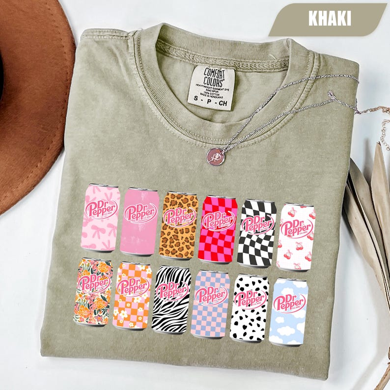 May include: A khaki crew neck t-shirt featuring a graphic print of twelve Dr. Pepper soda cans in various patterns including leopard print, checkerboard, zebra stripes, floral, and cow print. The shirt has a Comfort Colors brand label at the neck.