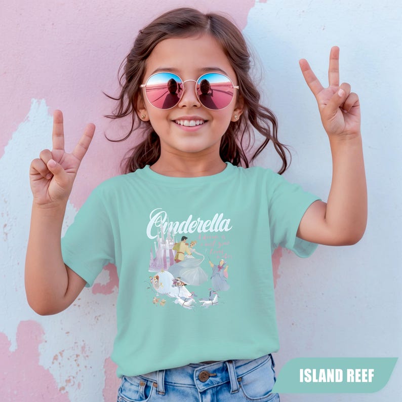 Peut inclure: Un t-shirt turquoise clair avec le mot "Cinderella" en &eacute;criture manuscrite. Le t-shirt pr&eacute;sente un graphisme de Cendrillon et de son ch&acirc;teau. La fille porte des lunettes de soleil rondes et fait un signe de paix.