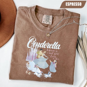 Peut inclure: T-shirt couleur expresso avec un motif Cendrillon. Le motif comprend le mot "Cendrillon" en &eacute;criture cursive, un ch&acirc;teau et des personnages du film. Un collier argent&eacute; avec un pendentif est drap&eacute; sur le t-shirt.