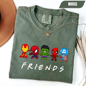 May include: Moss green t-shirt featuring cartoon illustrations of superheroes. The word "FRIENDS" is printed in white below the characters. A silver necklace with a pendant is draped on the shirt.