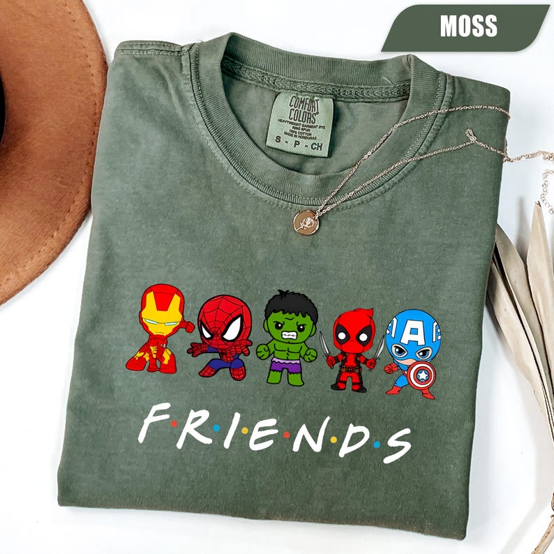 May include: Moss green t-shirt featuring cartoon illustrations of superheroes. The word "FRIENDS" is printed in white below the characters. A silver necklace with a pendant is draped on the shirt.