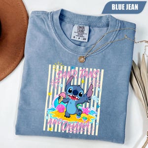 Op de afbeelding: Een blauw spijkerkleurig t-shirt met een afbeelding van het personage Stitch die een ijshoorntje vasthoudt op een zandstrand. Het ontwerp bevat de tekst SAND, TANS AND OHANA, palmbomen, sterren, een schelp, een zeester en een zonnebril.