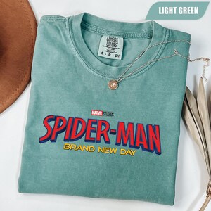 Może przedstawiać: Jasnozielona koszulka z logo Spider-Man z Marvel Studios w kolorze czerwonym i niebieskim oraz napisem "BRAND NEW DAY" w kolorze ż&oacute;łtym. Srebrny naszyjnik z wisiorkiem spoczywa na koszulce. Koszulka marki Comfort Colors.