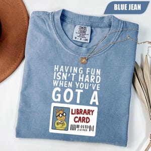 Op de afbeelding: Blauw spijkerbroekkleurig T-shirt met de tekst "HAVING FUN ISN'T HARD WHEN YOU'VE GOT A LIBRARY CARD." Voorzien van een afbeelding van Arthur de Aardvark en een bibliotheekkaart. Een gouden ketting ligt over het shirt.
