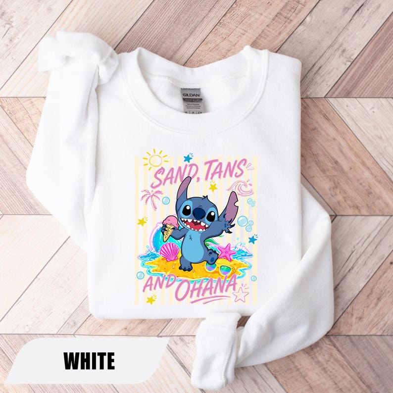 May include: A white crewneck sweatshirt featuring a graphic of the blue character Stitch holding an ice cream cone on a sandy beach. The design includes pink text that reads SAND, TANS AND OHANA, surrounded by sun, palm tree, and seashell icons.