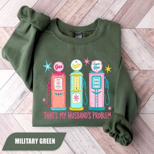 Pode incluir: Camisola verde militar com um gráfico de três bombas de gasolina vintage em rosa, azul e amarelo. O texto "That's My Husband's Problem" está abaixo das bombas. A camisola é feita de um material macio.