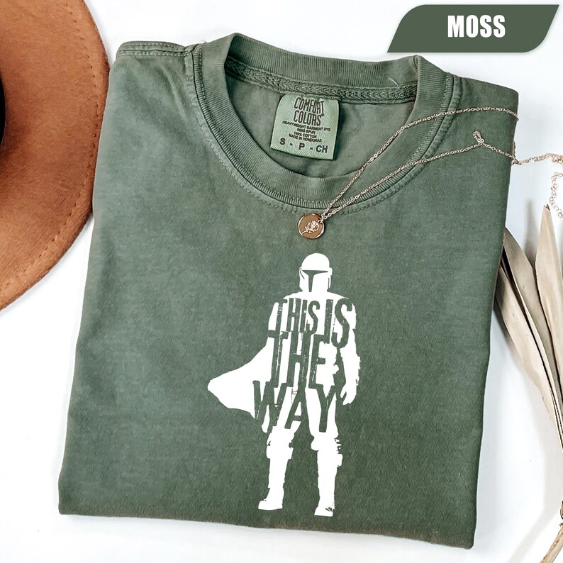 May include: A moss green short-sleeve t-shirt featuring a white graphic silhouette of a helmeted character with the text "THIS IS THE WAY" integrated into the design. The shirt has a Comfort Colors brand label inside the collar.