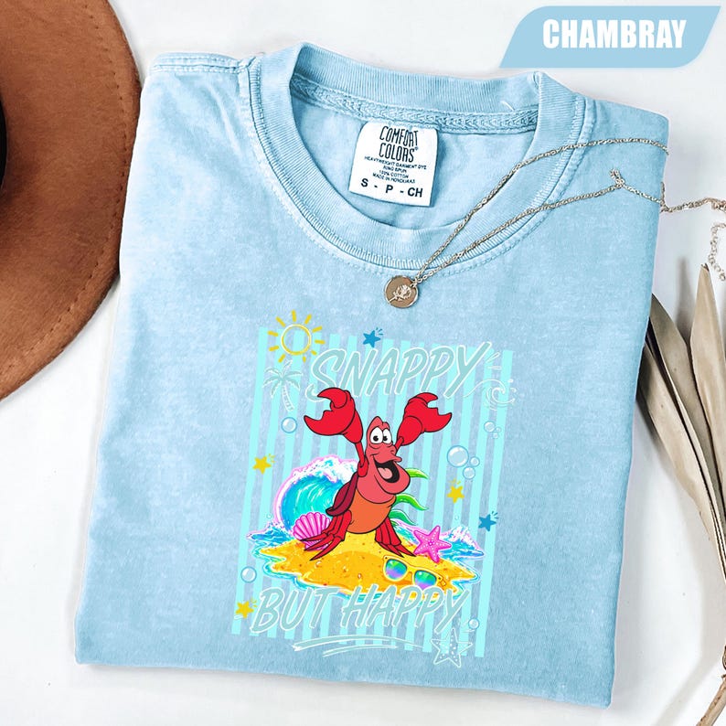 May include: Light blue t-shirt with a graphic of a red crab on a sandy beach with the words "Snappy But Happy". The shirt has a crew neck and short sleeves. A silver necklace is draped across the shirt.