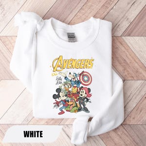 May include: A white crewneck sweatshirt featuring a graphic print of Disney characters dressed as Avengers. The design includes the text The Avengers, with illustrations of Mickey Mouse, Minnie Mouse, Goofy, Donald Duck, and Pluto, along with signatures.
