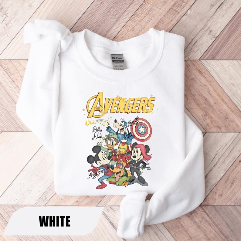 May include: A white crewneck sweatshirt featuring a graphic print of Disney characters dressed as Avengers. The design includes the text The Avengers, with illustrations of Mickey Mouse, Minnie Mouse, Goofy, Donald Duck, and Pluto, along with signatures.