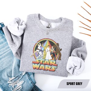 May include: A gray sweatshirt featuring a Star Wars graphic with characters and a rainbow design. The text "STAR WARS" is displayed in a colorful, retro font. The sweatshirt is paired with blue jeans.