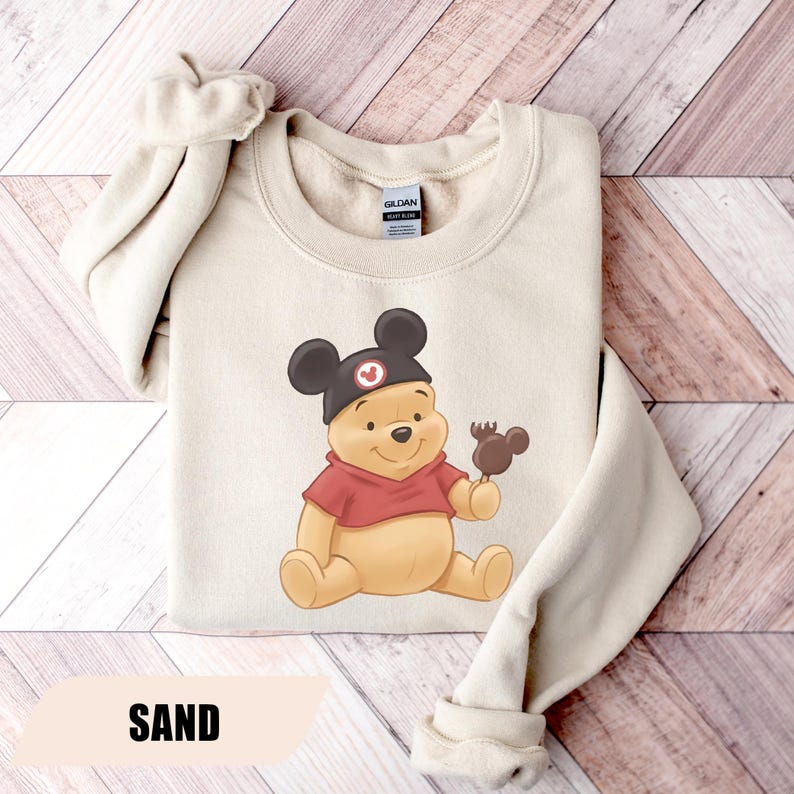 Pu&ograve; includere: Felpa color sabbia con un'illustrazione di Winnie the Pooh con un cappello di Topolino e che tiene in mano una caramella a forma di Mickey ricoperta di cioccolato. La felpa ha un girocollo e maniche lunghe. La parola "SAND" &egrave; stampata in basso.