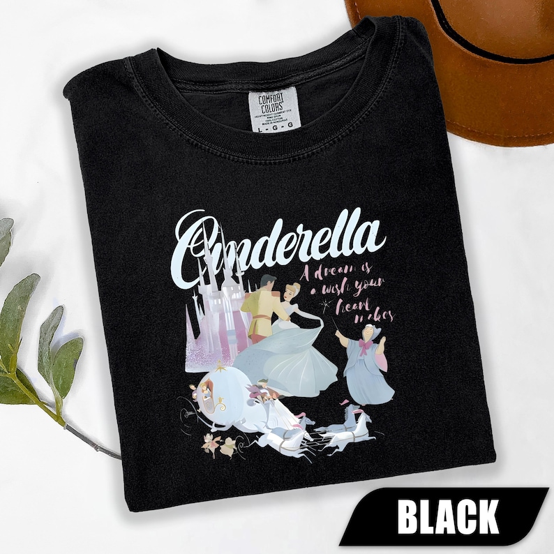 Peut inclure: T-shirt noir avec un motif Cendrillon. Le design comprend le mot "Cinderella" en cursive, un ch&acirc;teau et des illustrations de Cendrillon et d'autres personnages. Le texte "A dream is a wish your heart makes" est &eacute;galement inclus.