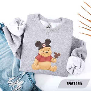 Pu&ograve; includere: Felpa grigio sport con un personaggio dei cartoni animati di Winnie the Pooh con un cappello di Topolino e che tiene in mano una leccornia al cioccolato. Il personaggio indossa una camicia rossa. La felpa ha un girocollo e maniche lunghe. Il testo "SPORT GREY" &egrave; in basso.