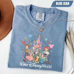 Puede incluir: Camiseta azul vaquero con un estampado de personajes de Toy Story frente a un castillo, con el texto "Walt Disney World". La camiseta tiene cuello redondo y mangas cortas. Un collar dorado est&aacute; drapeado sobre la camiseta.