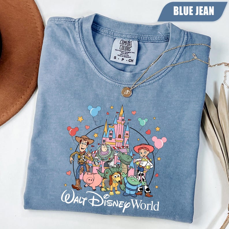 Puede incluir: Camiseta azul vaquero con un estampado de personajes de Toy Story frente a un castillo, con el texto "Walt Disney World". La camiseta tiene cuello redondo y mangas cortas. Un collar dorado est&aacute; drapeado sobre la camiseta.