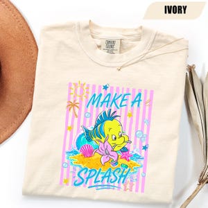 May include: Ivory-colored sweatshirt with a graphic of a yellow and blue cartoon fish on a small island with the words "MAKE A SPLASH". The design includes pink stripes, a sun, palm trees, and bubbles.