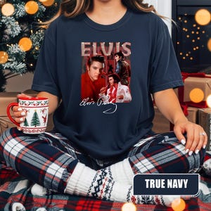 May include: A navy blue t-shirt featuring a graphic of Elvis Presley with the word "ELVIS" above the images. The shirt has a crew neck and short sleeves. The image includes a signature and multiple images of Elvis.