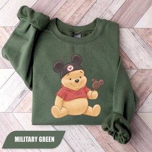 Pu&ograve; includere: Felpa verde militare con un'illustrazione di Winnie the Pooh con un cappello di Topolino e un gelato a forma di Mickey. La felpa ha maniche lunghe e girocollo. Il testo "MILITARY GREEN" &egrave; visualizzato su uno striscione verde.