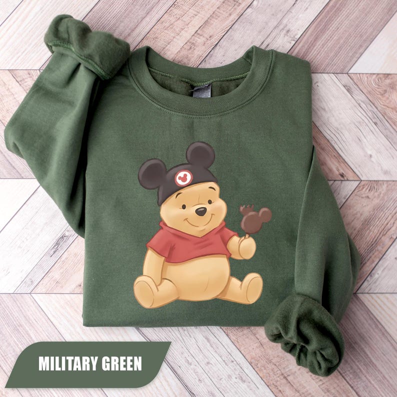 Pu&ograve; includere: Felpa verde militare con un'illustrazione di Winnie the Pooh con un cappello di Topolino e un gelato a forma di Mickey. La felpa ha maniche lunghe e girocollo. Il testo "MILITARY GREEN" &egrave; visualizzato su uno striscione verde.