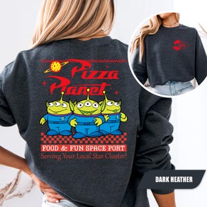Puede incluir: Sudadera gris oscuro con el logo de Pizza Planet y tres personajes alien&iacute;genas verdes con uniformes azules. La parte trasera de la sudadera tiene el texto "FOOD & FUN SPACE PORT Serving Your Local Star Cluster!" en rojo y blanco.