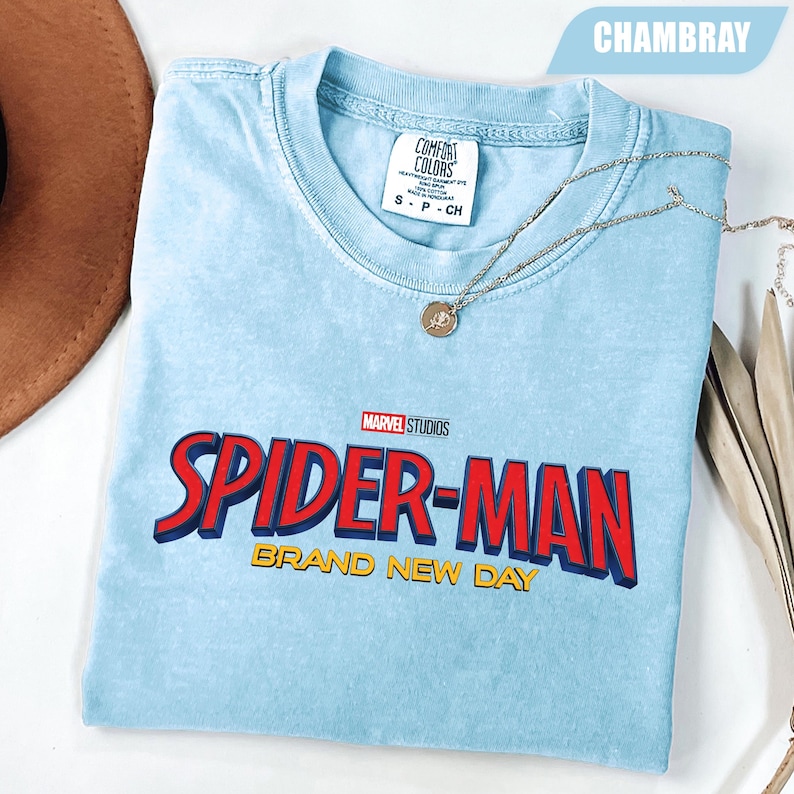 Może przedstawiać: Jasnoniebieska koszulka z logo Spider-Man Marvel Studios w kolorze czerwonym i niebieskim, z napisem "BRAND NEW DAY" w kolorze ż&oacute;łtym. Koszulka marki Comfort Colors. Srebrny naszyjnik z wisiorkiem jest na koszulce.