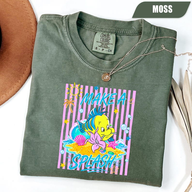 May include: Moss green t-shirt with a graphic featuring a yellow fish, pink and blue accents, and the words "MAKE A SPLASH". The shirt has a crew neck and short sleeves. A gold necklace is draped across the shirt.