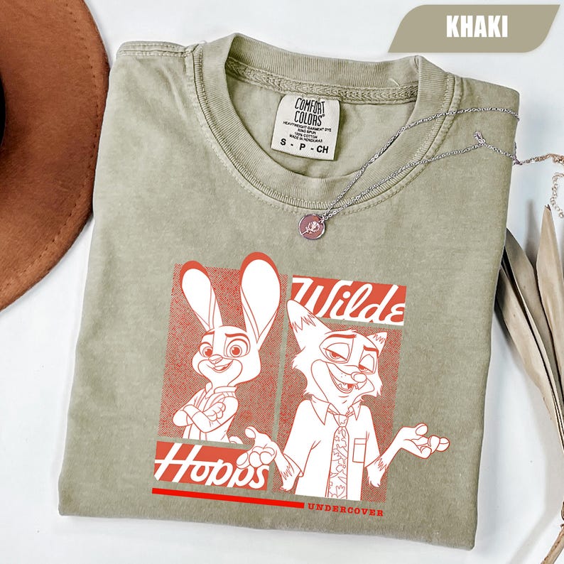 Puede incluir: Camiseta de color caqui con un gr&aacute;fico rojo y blanco de personajes de dibujos animados. El gr&aacute;fico incluye las palabras "Hopps" y "Wilde" con "Undercover" debajo. La camiseta est&aacute; hecha de un material suave y tiene una etiqueta Comfort Colors.