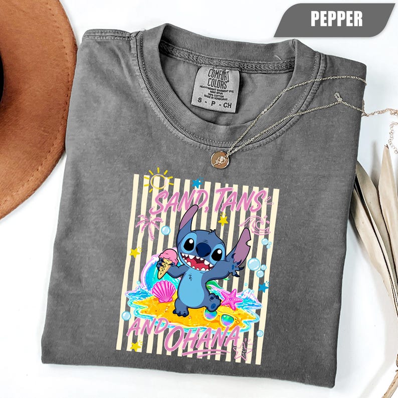 Op de afbeelding: Een antracietgrijs t-shirt met een afbeelding van het blauwe personage Stitch die een ijshoorntje vasthoudt. Het ontwerp bevat de tekst SAND, TANS AND OHANA, omringd door gele strepen, palmbomen, schelpen, sterren en bubbels.