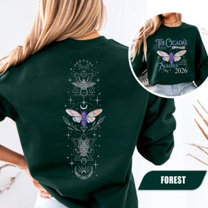 May include: Forest green sweatshirt featuring a detailed graphic of a cicada with celestial elements on the back. The front displays the text "The Cicada's Summer 2026" with a cicada illustration. The design includes geometric shapes and a crescent moon.