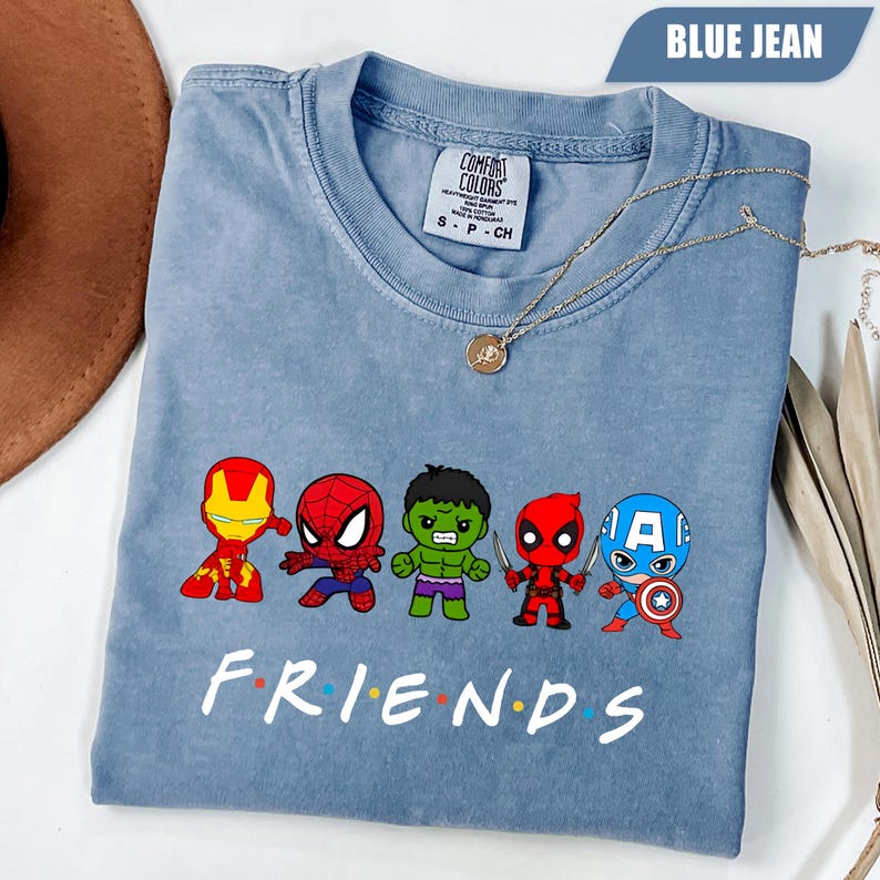 May include: Blue jean colored t-shirt featuring cartoon versions of Marvel characters Iron Man, Spider-Man, Hulk, Deadpool, and Captain America. The word "FRIENDS" is printed in white below the characters. A gold necklace is draped across the shirt.