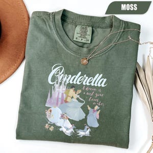 Peut inclure: T-shirt vert mousse avec un motif Cendrillon. Le motif pr&eacute;sente le mot "Cendrillon" en &eacute;criture cursive, un ch&acirc;teau et des illustrations de personnages. Le t-shirt porte &eacute;galement l'inscription "A dream is a wish your heart makes."