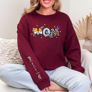 Op de afbeelding: Bordeaux sweatshirt met het woord "MOM" in kleurrijke, cartoon-stijl letters. De letter "M" heeft een personage, de "O" heeft een raket en de tweede "M" heeft een koeienprint. De linkermouw heeft stripfiguren.