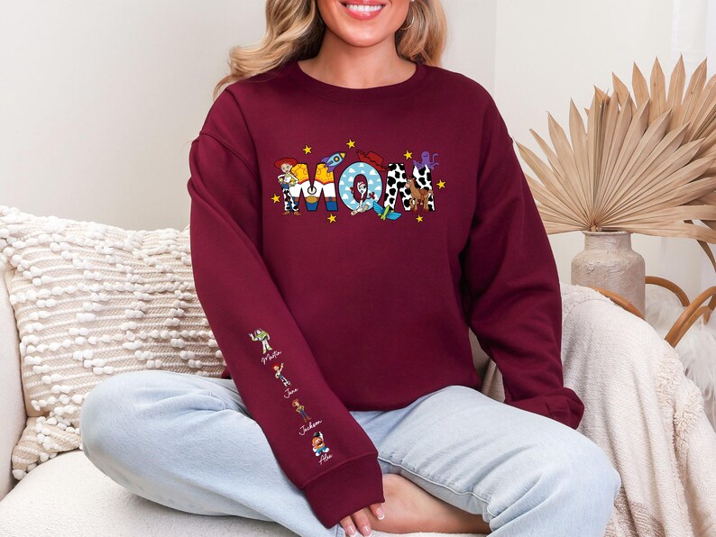 Op de afbeelding: Bordeaux sweatshirt met het woord "MOM" in kleurrijke, cartoon-stijl letters. De letter "M" heeft een personage, de "O" heeft een raket en de tweede "M" heeft een koeienprint. De linkermouw heeft stripfiguren.