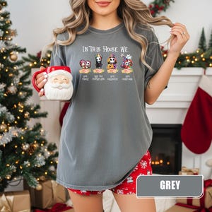 May include: A grey t-shirt with the text "IN THIS HOUSE WE" above cartoon characters. Below the characters are the words "LOVE FAMILY", "LOVE THE PUMPKIN KING", "LOVE HALLOWEEN", and "LOVE CHRISTMAS". The shirt is paired with red shorts.