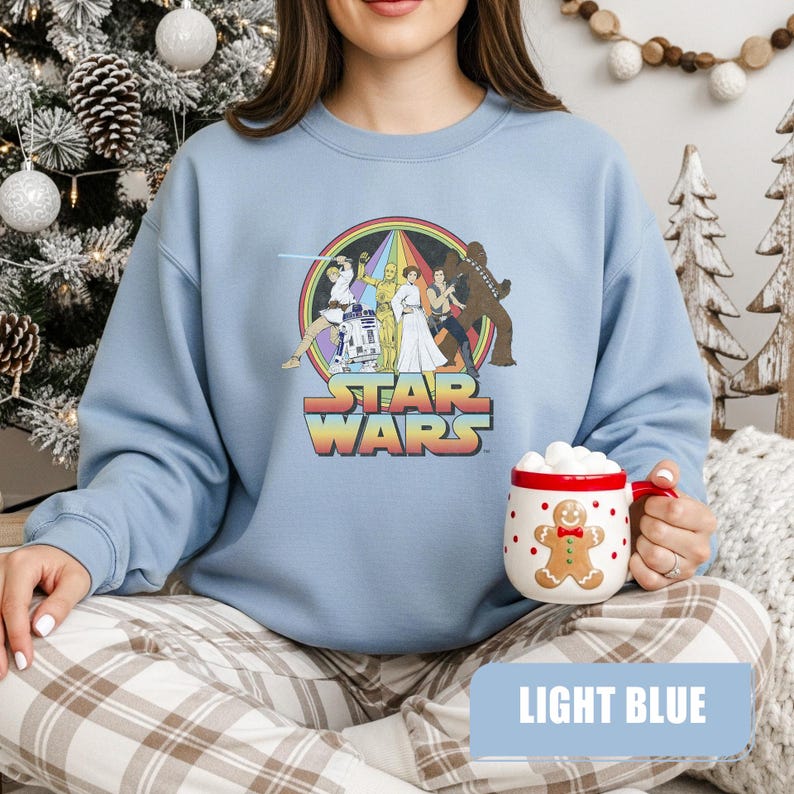 May include: Light blue Star Wars sweatshirt featuring a colorful graphic of characters from the movie. The sweatshirt has a crew neck and long sleeves. The text "STAR WARS" is displayed in a retro font. A gingerbread man mug is held in the hands.