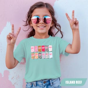 May include: A light mint green short-sleeve t-shirt featuring a graphic print of twelve soda cans arranged in two rows. Each can displays a unique pattern, including leopard print, zebra stripes, checkerboard, and floral designs, with the Dr Pepper logo.